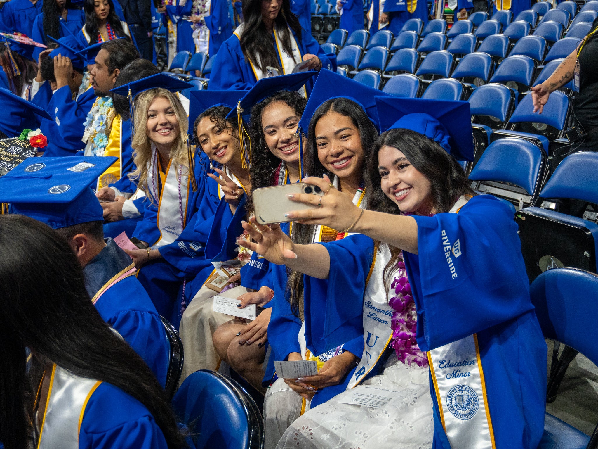 Five smiling CHASS graduates in blue caps and gowns sit in arena seating, leaning together to take a group selfie with a smartphone during a UC Riverside commencement ceremony.