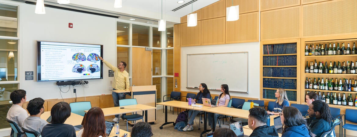 A presenter points to a brain scan diagram on a large monitor during a seminar with students seated around tables with laptops in a bright classroom.