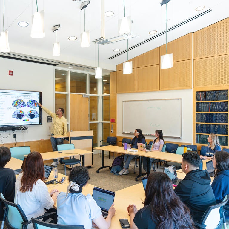 A presenter points to a brain scan diagram on a large monitor during a seminar with students seated around tables with laptops in a bright classroom.