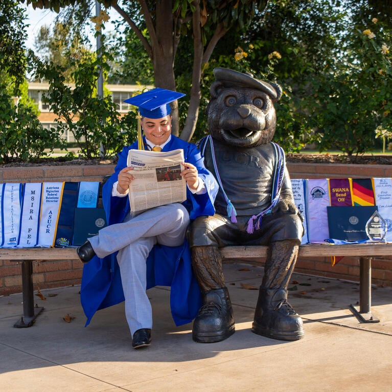 Antriana Bigiazian sits on a bench next to a bear statue, reading a newspaper in full blue graduation regalia with graduation sashes spread beside them. Photo by Lyndsey Rankin.