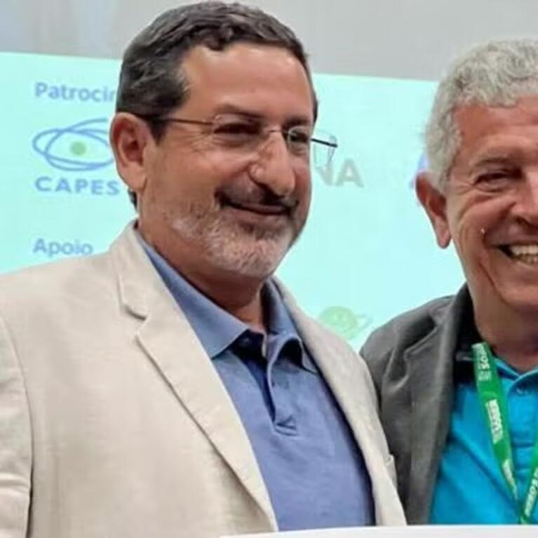 Steven Helfand (left), wearing glasses, a beige jacket, and a blue shirt, stands smiling next to another man in a gray jacket with a conference lanyard in front of a conference backdrop.