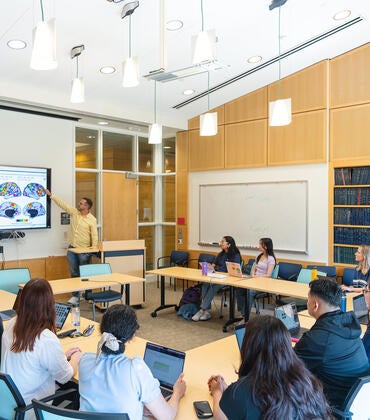 A presenter points to a brain scan diagram on a large monitor during a seminar with students seated around tables with laptops in a bright classroom.