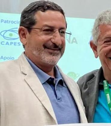 Steven Helfand (left), wearing glasses, a beige jacket, and a blue shirt, stands smiling next to another man in a gray jacket with a conference lanyard in front of a conference backdrop.