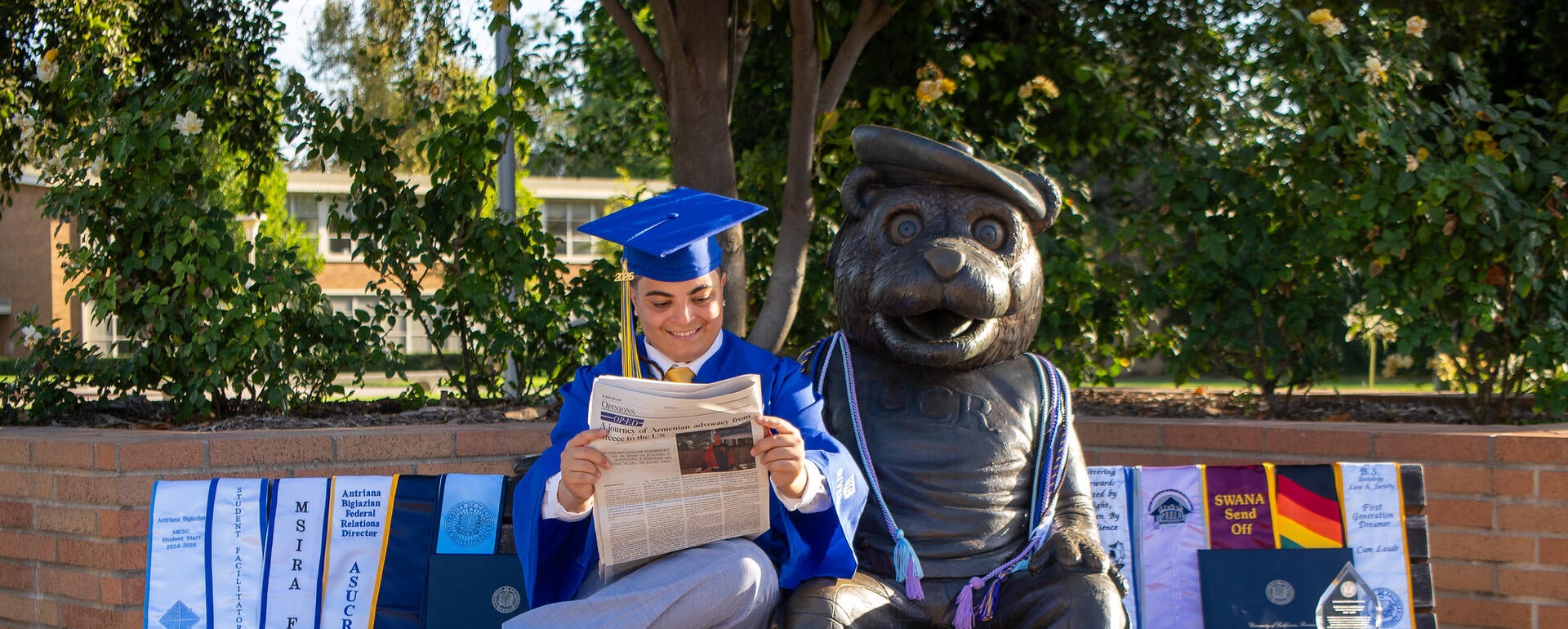 Antriana Bigiazian sits on a bench next to a bear statue, reading a newspaper in full blue graduation regalia with graduation sashes spread beside them. Photo by Lyndsey Rankin.