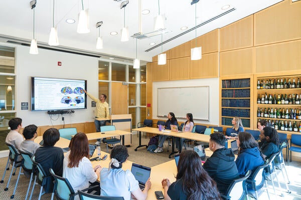 A presenter points to a brain scan diagram on a large monitor during a seminar with students seated around tables with laptops in a bright classroom.