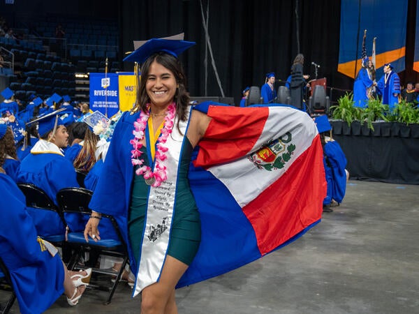 A smiling UCR CHASS graduate wears a blue cap, gown, pink lei, and custom sash while proudly holding open the flag of Peru inside an arena during a UC Riverside graduation ceremony.
