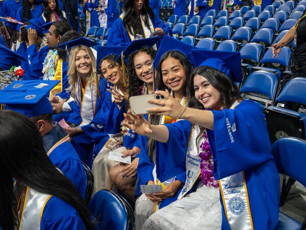 Five smiling CHASS graduates in blue caps and gowns sit in arena seating, leaning together to take a group selfie with a smartphone during a UC Riverside commencement ceremony.
