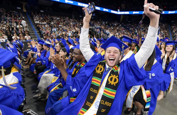 A UCR CHASS graduate wearing a blue cap, gown, and 'Class of 2026' stole cheers with arms raised high at a packed indoor graduation ceremony while surrounding classmates applaud.