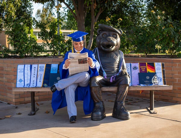 Antriana Bigiazian sits on a bench next to a bear statue, reading a newspaper in full blue graduation regalia with graduation sashes spread beside them. Photo by Lyndsey Rankin.