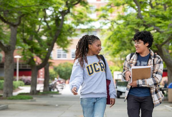 Two students walk together and chat on a leafy university campus pathway; one wears a gray UCR sweatshirt while the other carries notebooks.