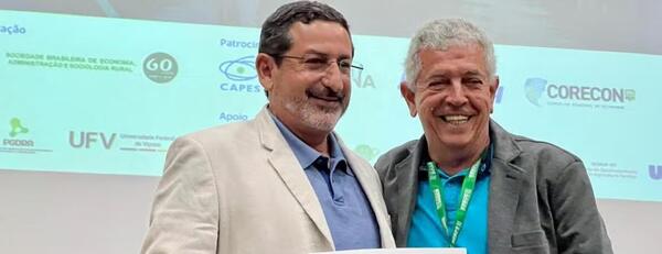 Steven Helfand (left), wearing glasses, a beige jacket, and a blue shirt, stands smiling next to another man in a gray jacket with a conference lanyard in front of a conference backdrop.