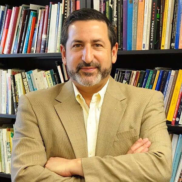Headshot of Steven Helfand with a trimmed beard, wearing a light tan jacket and collared shirt, posing with a slight smile in front of a bookshelf full of books.