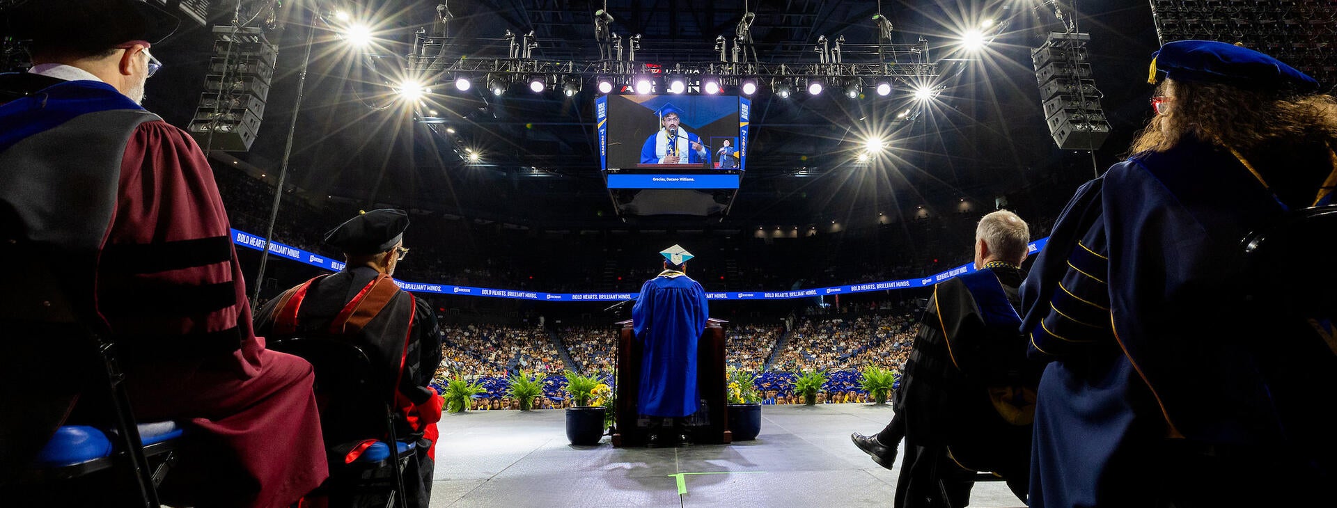  Looking from behind faculty on a stage, a graduate in a blue gown speaks at a podium in a crowded, dimly lit arena. A large overhead screen displays the speaker, framed by bright stage lights and banners reading "BOLD HEARTS. BRILLIANT MINDS."