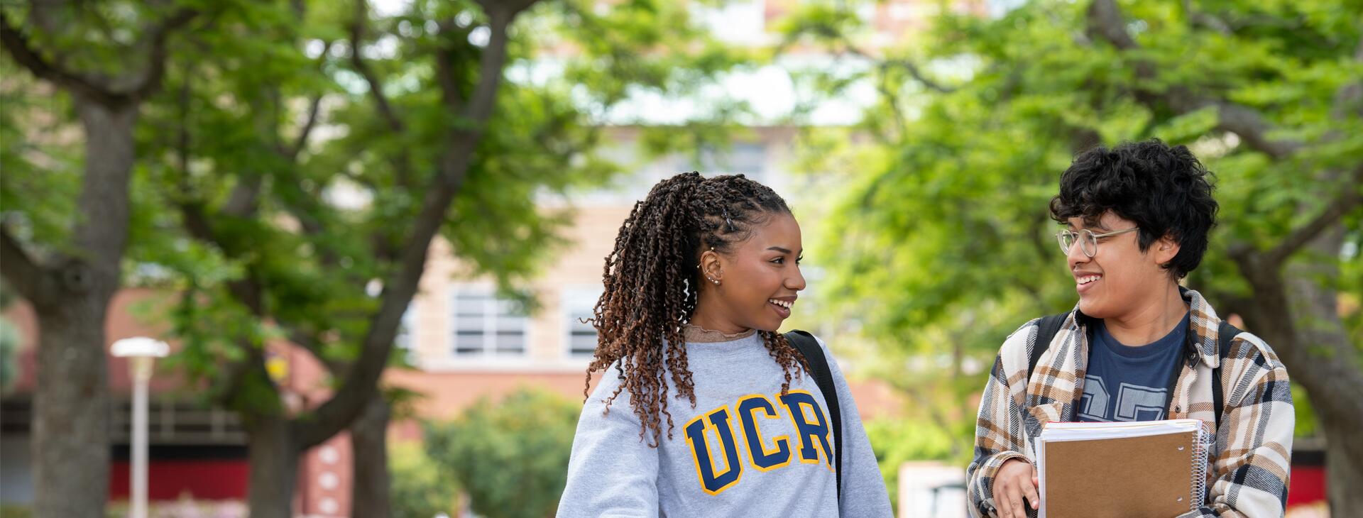 Two students walk together and chat on a leafy university campus pathway; one wears a gray UCR sweatshirt while the other carries notebooks.