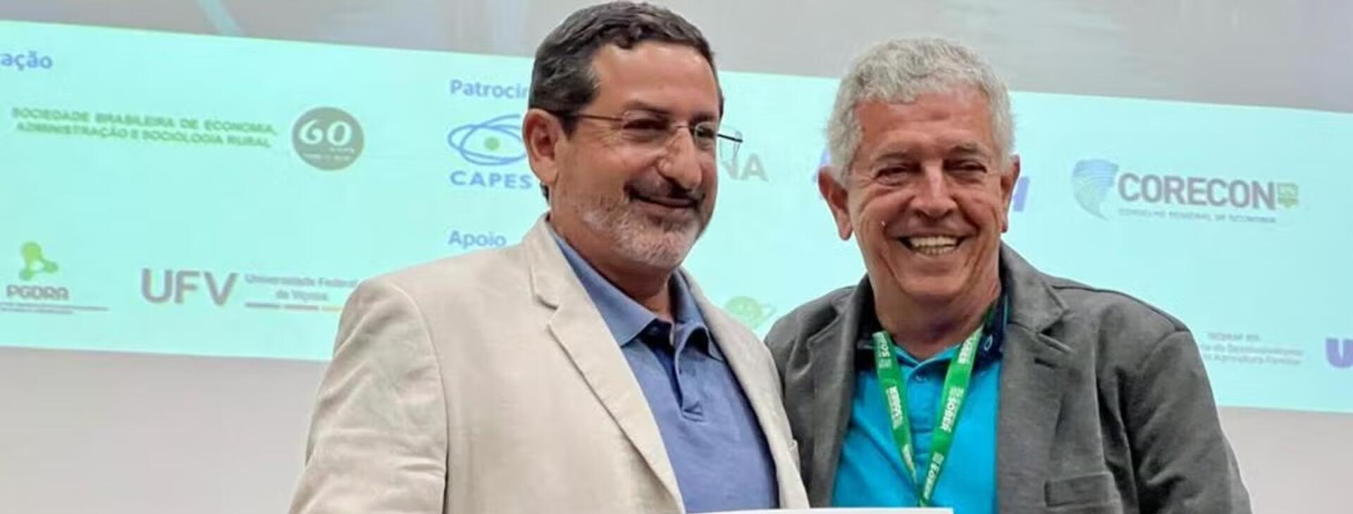 Steven Helfand (left), wearing glasses, a beige jacket, and a blue shirt, stands smiling next to another man in a gray jacket with a conference lanyard in front of a conference backdrop.